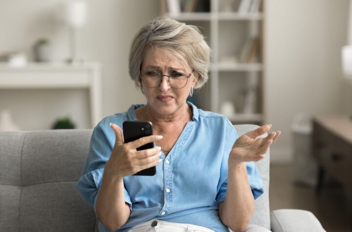 A mature woman in a blue blouse looking at an incoming phone call and wondering, “Is wireless caller spam?